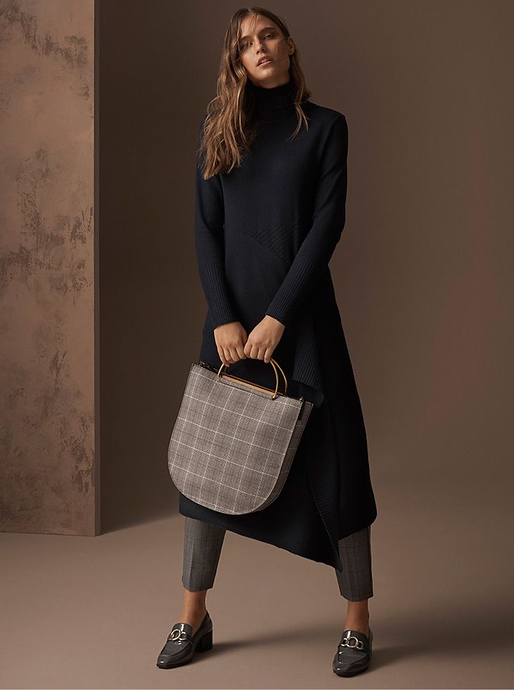 Model wears a navy jumper dress with an asymmetric hem over grey checked trousers with a grey checked top-handle bag and grey loafers