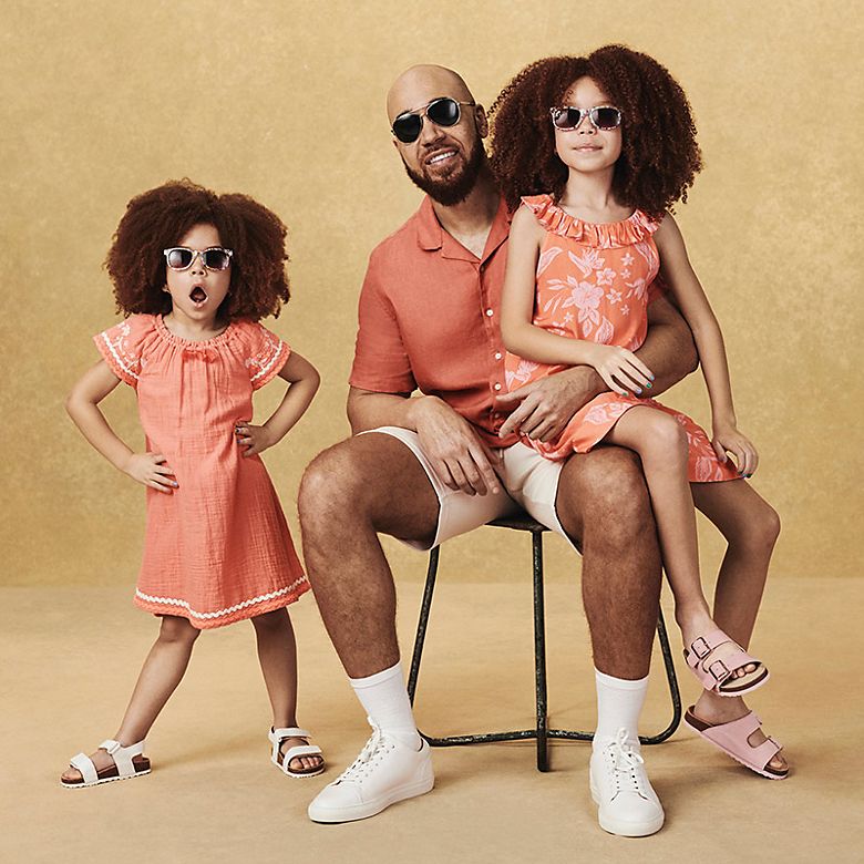 Man wearing orange shirt and beige shorts with his two young daughters wearing orange summer dresses