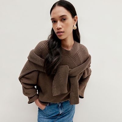 Woman wearing a mushroom brown jumper with a brown jumper knotted over her shoulders. Shop women’s knitwear 