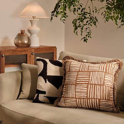 Room with beige sofa, graphic cushions, table lamp and wooden sideboards. Shop our homeware trends 