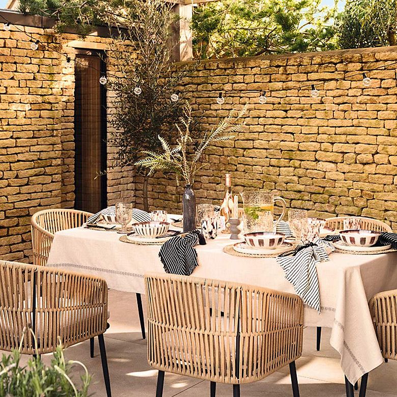 Outdoor dining table with rattan chairs set with a white tablecloth and crockery for al fresco dining. Shop outdoor furniture