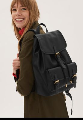 Woman wearing khaki trench coat, holding black buckled backpack