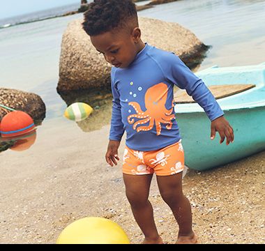 Boy wearing blue rash vest with octopus motif and matching orange shorts with yellow beach ball. Shop rash vests.