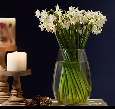 White narcissi bouquet in glass vase on sideboard with festive candles. Shop now