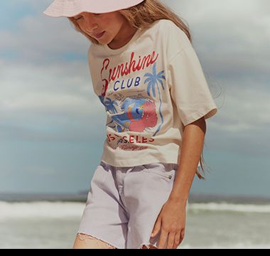 Girl wearing printed slogan T-shirt, shorts and a sunhat. Shop girls’ T-shirts