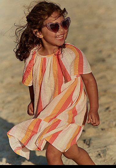 Girl wearing pink and orange striped summer dress. Shop girls’ dresses