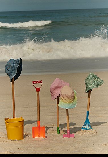 A collection of kids’ sun hats perched on spades in the sand. Shop girls’ sun hats