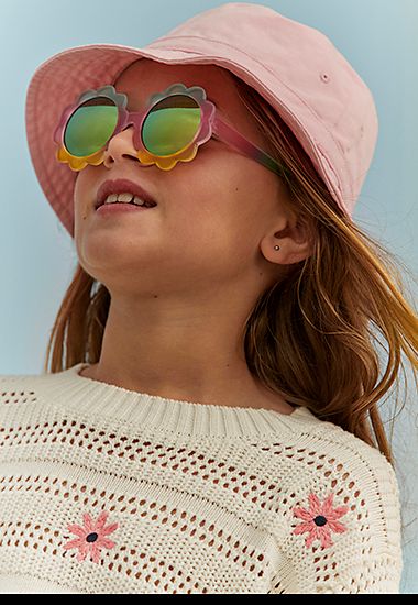 Girl wearing floral knitted top, sunglasses and pink sun hat. Shop girls’ holiday clothes