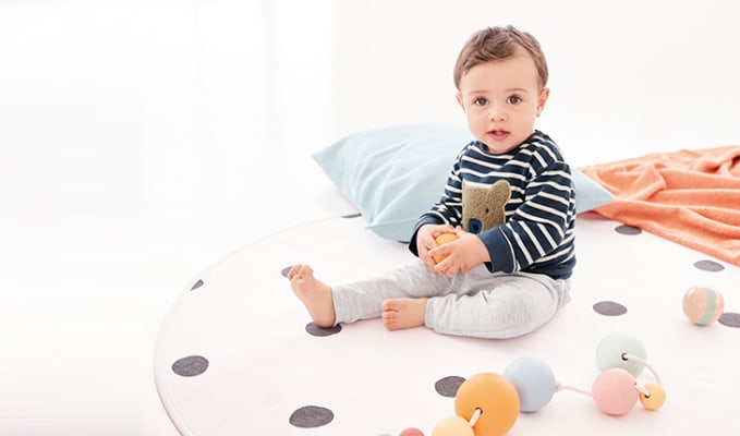 Baby Sat On A Playing Mat Wearing A Striped Teddy Bear Jumper And Grey Trousers